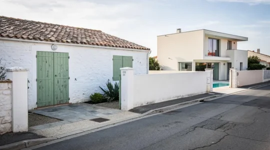 A whitewashed traditional cottage with green shutters stands on a quiet street near a modern glass-fronted villa on the Île de Ré.