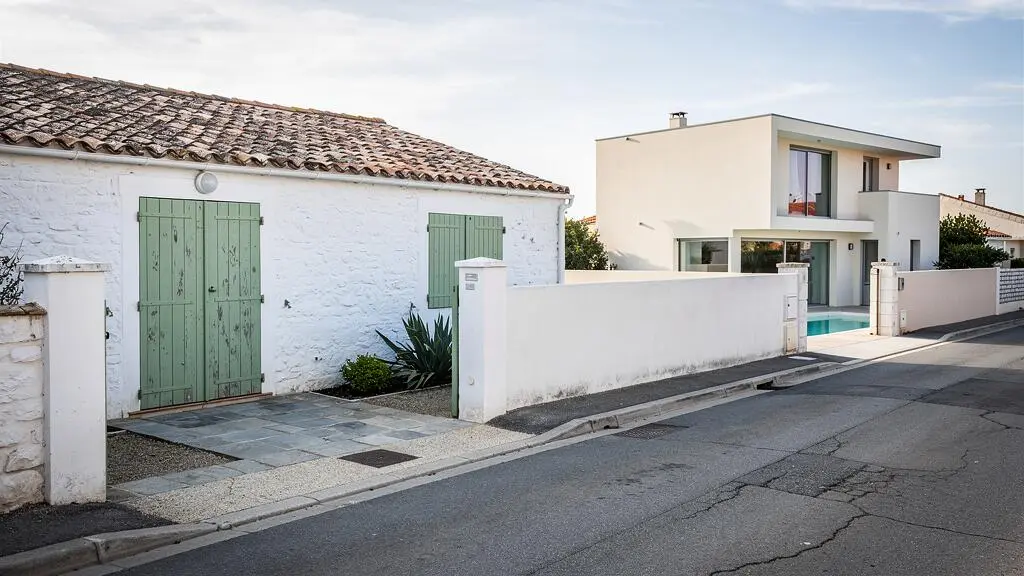 A whitewashed traditional cottage with green shutters stands on a quiet street near a modern glass-fronted villa on the Île de Ré.